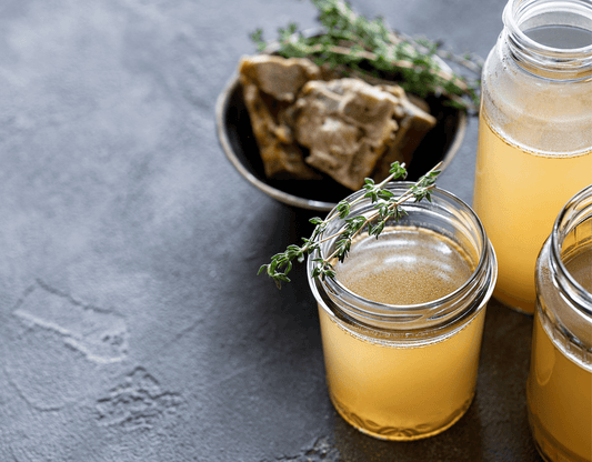 Glass jars of bone broth garnished with fresh thyme, surrounded by broth ingredients on a dark tabletop.